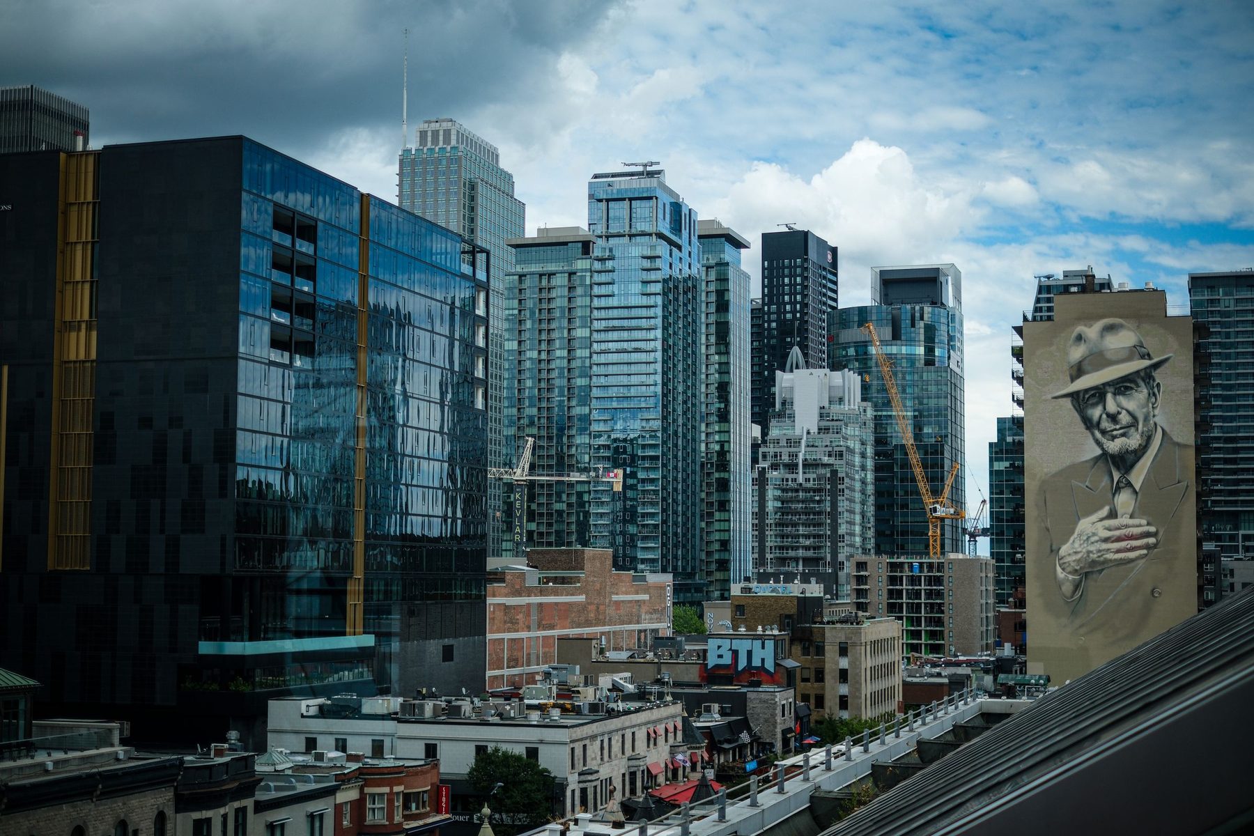 Downtown Montréal under a heavy sky — glass condo towers, cranes, and the Leonard Cohen mural on a building at the right.
