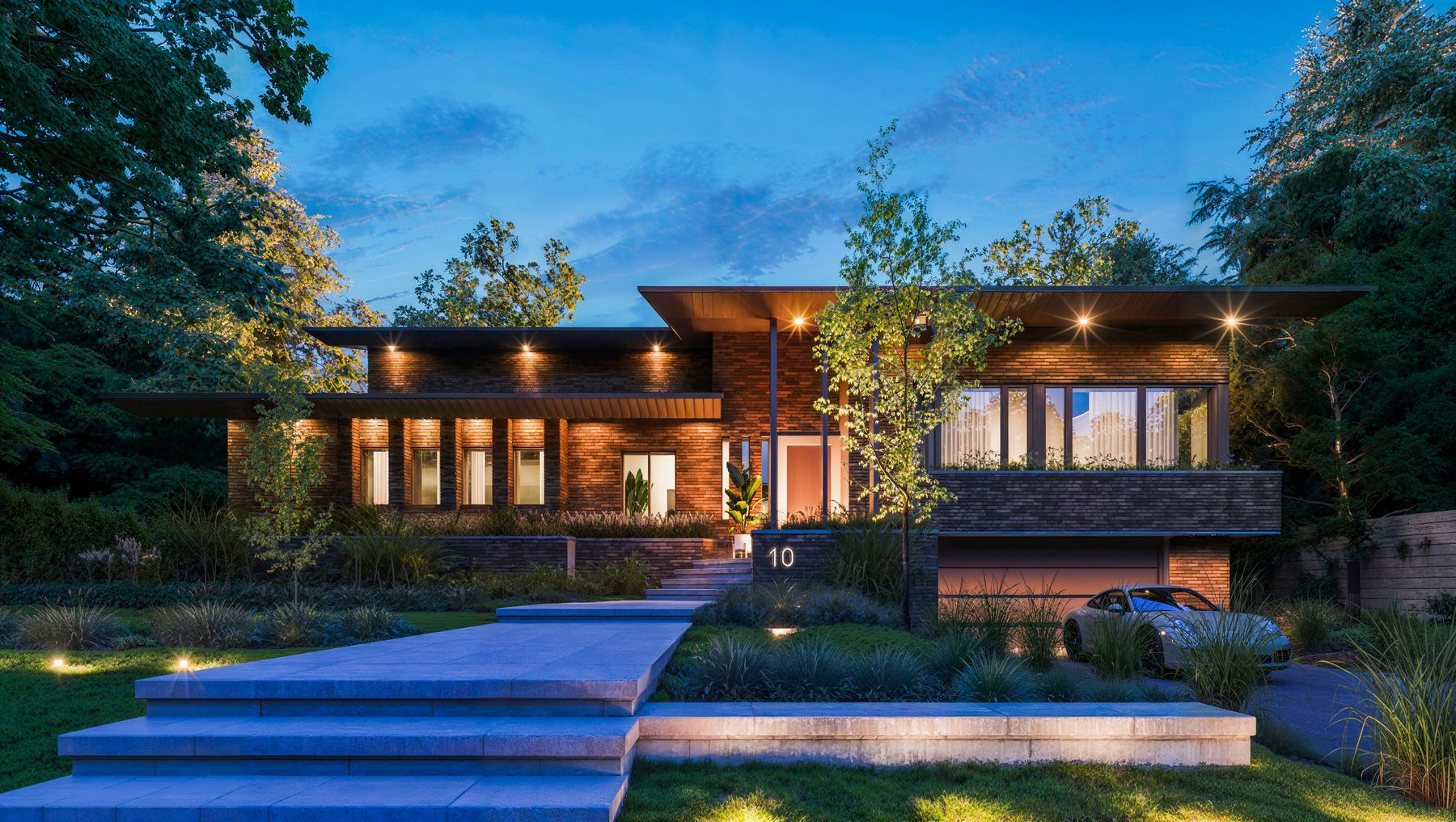 A contemporary Montréal house at dusk, its windows lit and its walk lined with planting, seen from the street.